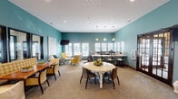 This single-floor community space is defined by its cohesive teal-painted walls and neutral carpet flooring, creating a calm and inviting atmosphere throughout. The main gathering area is thoughtfully divided into distinct functional zones that flow seamlessly into one another. A central lounge area features a mix of yellow upholstered armchairs and a light blue accent chair arranged around a rustic wood coffee table, complemented by a wall-mounted flat-screen television and abstract artwork. Adjacent to this is a dining area with a round white pedestal table and dark wood chairs, as well as a separate rectangular dining table with bench-style tufted seating and slipcover chairs. A row of dark-framed interior windows lines one wall, offering views into a fitness center beyond, effectively connecting the spaces visually. The kitchenette area is well-appointed with dark wood cabinetry, light granite countertops, a coffee station, microwave, and sink, bathed in natural light from multiple large windows with white blinds. Recessed lighting and pendant fixtures illuminate the space throughout, while high ceilings contribute to an open, airy feel. The overall design balances comfort and functionality with a consistent, polished aesthetic.