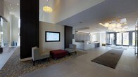 The lobby at 100 Danforth Avenue in Dobbs Ferry, New York, presents a polished and contemporary first impression for visitors and residents alike. The space is defined by clean architectural lines, a neutral color palette of grays and whites, and carefully curated design details that contribute to a professional yet welcoming atmosphere. Large-format tile flooring extends throughout, providing a seamless and refined foundation underfoot. A prominent reception desk with a textured white facade serves as the focal point of the space, positioned to greet all who enter. Adjacent to the entrance, a comfortable seating area features modern accent chairs and a rich red ottoman arranged atop a patterned area rug, offering a relaxed spot for waiting or conversation. A wall-mounted digital display panel provides building information and enhances the functionality of the lobby. Warm pendant lighting, including a sculptural floral-style fixture, adds visual interest and a welcoming glow against the high ceiling. Natural light filters in through large windows near the reception area, brightening the space and connecting the interior to the outdoors. Textured wall paneling in warm wood tones contrasts elegantly with darker architectural columns, adding depth and dimension to the overall design.