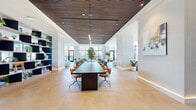 Located in Chicago's vibrant West Loop neighborhood, this generously proportioned single-floor amenity space at 225 N Elizabeth Street offers a sophisticated, open-plan environment designed for both collaboration and relaxation. The interior is defined by a seamless blend of modern design elements, including rich slatted wood ceiling panels that contrast beautifully against crisp white walls, and large-format stone tile flooring that runs throughout the entire space.

A central conference area features a long, dark-topped communal table surrounded by tan leather swivel chairs, anchored beneath a sleek linear pendant light. Flanking this zone is a striking navy blue open shelving unit, adorned with curated plants and decorative objects, which serves as a stylish divider between functional areas.

Toward the perimeter, floor-to-ceiling glass walls and oversized windows flood the space with abundant natural light while framing panoramic views of the surrounding Chicago cityscape. Multiple lounge zones are thoughtfully arranged throughout, furnished with a curated mix of leather, velvet, and upholstered seating in warm cognac, teal, and neutral tones. Lush potted trees and plants are positioned throughout, adding organic warmth to the contemporary palette. A game area and built-in cabinetry complete this versatile, amenity-rich environment.