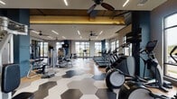 Located at 4015 Aspen Grove Dr in Franklin, TN, this single-floor fitness center is a well-appointed facility designed to support a comprehensive workout experience. The space is defined by its contemporary aesthetic, with striking hexagonal patterned flooring transitioning to warm hardwood in key areas, bold navy blue accent walls, and ceiling fans paired with sleek linear LED lighting throughout.

The main workout floor is generously proportioned, offering dedicated zones for both cardio and strength training. The cardio section features a row of treadmills, ellipticals, and a stair climber, all positioned along large floor-to-ceiling windows that flood the area with natural light and provide views of the surrounding landscape. The strength training zone includes a full array of cable and resistance machines, a dumbbell rack, and a spacious functional training area outfitted with a power rack, battle ropes, and plyo boxes — all set on durable rubberized flooring. Large mirrors line the walls to maximize the sense of space.

A unique feature is the indoor-outdoor workout area, accessible via a glass garage door that opens directly onto a turf lawn, seamlessly blending interior and exterior training spaces. This area provides additional equipment storage and a versatile setting for group or open-air exercise. The overall ambiance is energetic yet refined, making this fitness center a standout amenity.