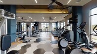Located at 4015 Aspen Grove Dr in Franklin, TN, this single-floor fitness center is a well-appointed facility designed to support a comprehensive workout experience. The space is defined by its contemporary aesthetic, with striking hexagonal patterned flooring transitioning to warm hardwood in key areas, bold navy blue accent walls, and ceiling fans paired with sleek linear LED lighting throughout.

The main workout floor is generously proportioned, offering dedicated zones for both cardio and strength training. The cardio section features a row of treadmills, ellipticals, and a stair climber, all positioned along large floor-to-ceiling windows that flood the area with natural light and provide views of the surrounding landscape. The strength training zone includes a full array of cable and resistance machines, a dumbbell rack, and a spacious functional training area outfitted with a power rack, battle ropes, and plyo boxes — all set on durable rubberized flooring. Large mirrors line the walls to maximize the sense of space.

A unique feature is the indoor-outdoor workout area, accessible via a glass garage door that opens directly onto a turf lawn, seamlessly blending interior and exterior training spaces. This area provides additional equipment storage and a versatile setting for group or open-air exercise. The overall ambiance is energetic yet refined, making this fitness center a standout amenity.