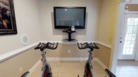 Located at 5001 Riverchase Drive in Phenix City, Alabama, this single-floor fitness space is thoughtfully designed to support an active lifestyle. The room is finished with neutral, warm-toned walls accented by clean white crown molding and baseboards, lending a polished and well-maintained appearance throughout.

The space is equipped with multiple stationary cycling bikes positioned directly facing a large wall-mounted flat-screen television, supported by a sleek wall-mounted media shelf — an ideal setup for engaging, entertainment-driven workouts. Light-toned tile flooring runs throughout the room, offering durability and easy maintenance appropriate for a high-use fitness environment.

A large mirror is mounted along one wall, visually expanding the space and providing functional feedback during exercise. Additional fitness equipment, including a treadmill and other strength or cardio machines, can be seen positioned throughout the room, making this a well-rounded, multi-use workout facility.

Natural light filters in through a window, brightening the interior and contributing to an energizing atmosphere. The overall layout is clean, organized, and purpose-built, making this space a fully functional private fitness center suited for a variety of training routines.