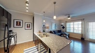 Located at 8823 Dugas Rd in San Antonio, TX, this single-floor residence offers a thoughtfully designed open-concept layout that seamlessly connects the kitchen, dining, and living areas. The kitchen is a standout feature, highlighted by a curved granite-topped island with a double-basin sink, elegant pendant lighting, track lighting overhead, and dark espresso cabinetry paired with black appliances. Crown molding adds a refined finishing touch throughout the common areas. The living room is warm and inviting, featuring rich hardwood-style flooring, crown molding, a ceiling fan, and large windows with exterior door access that flood the space with natural light and offer views to the outdoors. The bedroom is a comfortable retreat, complete with its own ceiling fan, hardwood-style flooring, a generous closet, and a window that brings in soft natural light. The bathroom features dark cabinetry with a granite-topped vanity, a large mirror framed by overhead lighting, and consistent hardwood-style flooring that carries through from the rest of the unit. Throughout the residence, a neutral color palette and cohesive design elements create a modern yet comfortable ambiance ideal for everyday living.