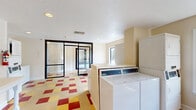 Located at 501 16th Ave N in Phenix City, Alabama, this shared laundry facility offers a functional and well-organized space designed to serve its users efficiently. The single-floor layout makes practical use of its compact footprint, accommodating multiple coin-operated washing machines and stacked dryer units positioned along the walls for an organized workflow.

The space is immediately distinctive thanks to its bold, multicolored checkered vinyl tile flooring in shades of red, yellow, and cream — a retro-inspired design element that adds visual energy to an otherwise utilitarian environment. Walls are finished in a neutral beige, with one accent wall painted a rich red, contributing to the room's lively character.

A white folding table with decorative turned legs runs along one wall, providing a dedicated surface for sorting and folding laundry. Framed informational notices are mounted nearby for resident convenience. A fire extinguisher and security camera are also visible, reflecting attention to safety.

Natural light enters through multiple large windows that offer views of the surrounding greenery and exterior grounds, keeping the space feeling open and airy. A set of glass-panel entry doors with a black frame connects the laundry room to a vestibule, allowing easy access. Recessed ceiling lighting and standard HVAC vents complete the practical, no-frills design of this well-maintained shared amenity space.