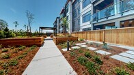 Located at 7 Erskine Lane in Victoria, BC, this thoughtfully designed outdoor communal space serves as the landscaped courtyard of a contemporary multi-storey residential building. The architecture of the building itself is modern and striking, featuring a grey and white panelled exterior, large windows, and glass-railed balconies that overlook the grounds below.

The centerpiece of the courtyard is a wide, smooth concrete walkway that runs the length of the space, flanked on both sides by generously sized raised garden beds constructed from warm cedar timber. These planters are filled with a variety of young plants and ground cover, set against a backdrop of red mulch that gives the landscape a rich, earthy tone. Slender bollard lighting is interspersed throughout, providing subtle illumination along the path.

A dark-framed pergola sits at the far end of the walkway, offering a shaded gathering area that complements the natural aesthetic of the grounds. Young trees have been planted throughout the space, adding vertical greenery that will mature over time and further enhance the ambiance.

Cedar fencing defines the perimeter of the courtyard, creating a sense of privacy and enclosure while maintaining a warm, natural character. The space enjoys abundant natural light and open sky views, making it an inviting and functional amenity for residents. The overall design reflects a strong commitment to community-oriented, sustainable outdoor living.