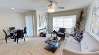 This single-floor space features an open-concept layout that seamlessly connects the living and dining areas, creating a cohesive and functional environment. The living area is anchored by plush wall-to-wall carpeting and centered around a dark-finished coffee table atop a woven area rug, complemented by a neutral-toned sofa and an upholstered accent chair. A ceiling fan with an integrated light fixture is centrally mounted, providing both airflow and ambient lighting. A large window fitted with horizontal blinds allows generous natural light to fill the room while offering adjustable privacy. A sliding door with vertical blinds leads to an adjacent area, likely an outdoor space. A sleek console table against the wall serves as a decorative focal point, paired with colorful abstract wall art that adds visual interest. The dining area, partially visible, includes dark dining chairs that coordinate with the overall color palette. Neutral wall tones and light-colored ceilings contribute to a clean, airy ambiance throughout. Tasteful greenery and decorative accents complete the well-balanced, inviting atmosphere of this thoughtfully arranged space.