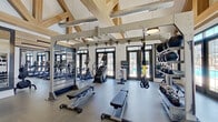Located at 5206 Wurzbach Pkwy in San Antonio, Texas, this fitness center is a standout amenity defined by its striking architectural character and comprehensive equipment offerings. The space is anchored by dramatic vaulted ceilings with exposed wood trusses and industrial-style pendant lighting, creating an open, airy atmosphere that feels both refined and functional. Large black-framed French doors and transom windows line the walls, flooding the interior with natural light and offering views of an outdoor pool area. The flooring transitions between durable rubber and wood-look surfaces, accommodating a wide variety of workout zones. The main floor is generously appointed with cardio equipment including treadmills, ellipticals, and a stair climber, alongside a robust RAX functional training rig, TRX suspension systems, weight benches, cable machines, and a full rack of dumbbells. A dedicated studio space is separated by floor-to-ceiling glass panels and mirrors, ideal for stretching, yoga, or group fitness. Decorative touches such as an abstract wall mural and a neatly organized towel station add to the polished, welcoming environment. A secondary exit door provides convenient outdoor access. Overall, the fitness center combines architectural elegance with practical, high-quality amenities to deliver an exceptional workout experience.