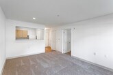 This single-floor residence offers a straightforward, functional layout designed for comfortable everyday living. The living area is generously proportioned, finished with neutral-toned wall-to-wall carpeting and smooth white walls that create a clean, bright atmosphere. A distinctive pass-through window connects the living space to the kitchen, enhancing the open feel between the two areas. Recessed lighting provides consistent illumination throughout.

The kitchen is efficiently arranged in a U-shaped configuration, featuring light wood cabinetry that extends to the ceiling for ample storage. White appliances, including an electric range with overhead ventilation, a dishwasher, and a refrigerator, are complemented by light-toned countertops and tile flooring, maintaining a cohesive, practical aesthetic.

The bedroom is a well-sized, carpeted space featuring two windows that frame views of surrounding greenery, allowing natural light to fill the room. An adjacent bedroom or secondary room also benefits from a window overlooking the outdoors.

The bathroom includes a single-sink vanity with light wood cabinetry, a large mirror with overhead lighting, and tile flooring consistent with the kitchen. The overall ambiance of the space is neutral, understated, and move-in ready.