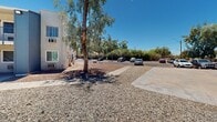 Located at 8028 S Central Ave in Phoenix, Arizona, this single-story and two-story residential complex offers a well-established community setting in the heart of the Sonoran Desert. The exterior grounds are finished with low-maintenance desert landscaping, featuring gravel terrain accented by mature eucalyptus, olive, and palm trees that provide natural shade throughout the property.

The complex is organized around a central courtyard that serves as the communal hub for residents. A fenced swimming pool is visible alongside towering palm trees, offering a classic Arizona outdoor retreat. Adjacent to the pool area, a basketball court provides an additional recreational outlet for residents.

The residential buildings themselves feature a practical two-story design with exterior staircases, individual unit entrances, and wall-mounted air conditioning units suited to the desert climate. The façade is finished in neutral tones with blue accent doors and railings, lending a cohesive and tidy appearance.

On-site parking is readily available, with a dedicated lot bordered by mature trees and gravel ground cover. The property's desert-adapted landscaping minimizes water usage while maintaining an inviting outdoor environment. Situated in South Phoenix, the complex enjoys easy access to Central Avenue and the surrounding neighborhood amenities.