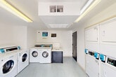 This shared laundry facility is a single-room space designed for efficient, practical use. The room features white walls, a textured ceiling with fluorescent overhead lighting, and durable speckled vinyl flooring that is easy to maintain. A glass-paneled door on one side of the room allows natural light to filter in while providing a direct exterior access point.

The laundry room is well-equipped with a row of front-loading commercial washers along one wall, complemented by a bank of stacked commercial dryers on the opposing wall — offering ample capacity for multiple users. Each machine is fitted with a digital card reader, and signage throughout the room promotes app-based payment, adding a modern, convenient touch to the facility. A narrow folding or sorting shelf runs above the washers, providing a functional surface for laundry tasks.

The layout is straightforward and uncluttered, maximizing the usable floor space within the room. A secondary solid door on another wall provides an additional entry or exit point. The overall ambiance is clean, bright, and utilitarian, reflecting a well-maintained shared amenity space purpose-built to serve the laundry needs of its users efficiently.