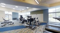 This fitness center offers a spacious, single-floor layout designed to accommodate a comprehensive range of workout activities. The space features high ceilings with recessed panel lighting and a drop ceiling that contributes to an open, airy atmosphere. Large windows along one wall allow an abundance of natural light to fill the room and provide views of the surrounding outdoor greenery, creating an inviting environment for exercise.

The flooring is a combination of patterned carpet and protective rubber mats positioned strategically beneath the equipment, offering both comfort and safety. The walls are painted in a neutral palette accented with muted blue and green tones, giving the space a calm, modern aesthetic.

The cardio section is well-stocked with a variety of machines, including stationary bikes, ellipticals, and other aerobic equipment arranged to allow ample circulation throughout the floor. A dedicated strength training area features a dumbbell rack and weight benches, offering options for resistance and free weight workouts. The overall layout is thoughtfully organized to clearly define zones for different types of exercise while maintaining an open, uncluttered feel throughout the space.