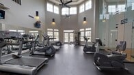 This fitness center offers a comprehensive workout environment designed to accommodate a wide range of exercise needs. The main gym area is defined by dramatically soaring ceilings that create an open, airy atmosphere, amplified by rows of large windows along the walls and French doors that flood the space with natural light and provide views of the surrounding exterior. A ceiling fan anchors the upper space, contributing to airflow and comfort during workouts.

The gym floor is finished with dark rubber flooring, providing durability and traction throughout the space. Equipment includes multiple treadmills, elliptical trainers, a stationary bike, and a multi-station cable strength machine, offering a well-rounded selection for both cardio and resistance training. A dedicated free weights rack with dumbbells is positioned along one wall. Full-length mirrors line portions of the walls, visually expanding the space while allowing users to monitor their form. Multiple wall-mounted televisions are positioned for easy viewing from the cardio equipment.

Adjacent to the main fitness area is a dedicated racquetball court, featuring a classic hardwood floor with regulation court markings and crisp white walls. Overhead fluorescent lighting ensures the court is well-illuminated for active play, rounding out this facility's diverse athletic offerings.