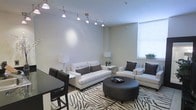 This single-floor space presents a well-appointed living area designed with a contemporary aesthetic. The room features notably high ceilings that enhance the sense of openness, complemented by a large window fitted with horizontal blinds that allows generous natural light to fill the space. A sleek track lighting system runs across the ceiling, providing adjustable, directed illumination throughout the room.

The living area is anchored by a white upholstered sofa and a matching sectional or accent chair, both accented with dark brown throw pillows that create a striking contrast. A bold zebra-print area rug defines the seating zone and adds visual interest to the neutral-toned flooring. At the center of the arrangement sits a round, dark leather ottoman that doubles as a coffee table, topped with a decorative tray.

Soft ambient lighting is provided by two matching table lamps positioned at either end of the room. A small side table holds a simple floral arrangement, adding a subtle organic touch. Adjacent to the living area, a dark dining table with matching chairs is partially visible, suggesting a seamless open-plan layout. The overall color scheme of whites, beiges, and dark browns contributes to a cohesive, modern ambiance.