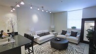 This single-floor space presents a well-appointed living area designed with a contemporary aesthetic. The room features notably high ceilings that enhance the sense of openness, complemented by a large window fitted with horizontal blinds that allows generous natural light to fill the space. A sleek track lighting system runs across the ceiling, providing adjustable, directed illumination throughout the room.

The living area is anchored by a white upholstered sofa and a matching sectional or accent chair, both accented with dark brown throw pillows that create a striking contrast. A bold zebra-print area rug defines the seating zone and adds visual interest to the neutral-toned flooring. At the center of the arrangement sits a round, dark leather ottoman that doubles as a coffee table, topped with a decorative tray.

Soft ambient lighting is provided by two matching table lamps positioned at either end of the room. A small side table holds a simple floral arrangement, adding a subtle organic touch. Adjacent to the living area, a dark dining table with matching chairs is partially visible, suggesting a seamless open-plan layout. The overall color scheme of whites, beiges, and dark browns contributes to a cohesive, modern ambiance.