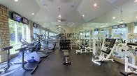 This fitness center is a thoughtfully designed, single-floor facility built to support a full range of workout routines. The space is defined by its striking vaulted ceiling, which lends an open, airy feel and is complemented by a combination of recessed lighting and hanging pendant-style ceiling fans that ensure the room stays bright and comfortable during any workout. Large windows along the perimeter walls flood the space with natural light, while stone accent columns add a distinctive architectural character.

Floor-to-ceiling mirrors line the walls, visually expanding the space and providing exercisers with proper form feedback from any position. The dark rubberized flooring offers both durability and safety across the entire workout area.

On the cardio side, a generous row of elliptical trainers and additional cardio machines are neatly arranged along one wall, offering users a variety of low- and high-impact options. At the center of the room, a well-stocked dumbbell rack provides a comprehensive range of free weights, accompanied by a dedicated workout bench. The strength training section features multiple cable and weight machines, supporting a broad array of resistance exercises. Mounted televisions are positioned for easy viewing, enhancing the overall workout experience in this well-appointed facility.