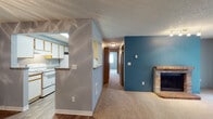 This single-floor unit is designed with a practical and comfortable layout. The living area serves as the heart of the space, highlighted by a striking brick fireplace set against a bold teal accent wall. Soft carpet underfoot adds warmth, while a sliding glass door and adjacent window allow natural light to fill the room and provide outdoor views. A ceiling fan adds functionality to the open living and dining area, which connects seamlessly to the kitchen via a pass-through window. The galley-style kitchen is efficiently arranged with white cabinetry trimmed in warm wood tones, laminate countertops, wood-style vinyl flooring, and white appliances including a range, refrigerator, and dishwasher. A skylight brightens the kitchen further. A hallway leads to the bedrooms, each finished with neutral carpet and soft gray walls. One bedroom features a window with views of surrounding greenery and a baseboard heater. The bathrooms feature wood-style flooring, laminate countertops, white cabinetry with wood-tone frames, and well-lit vanity mirrors. Textured ceilings are consistent throughout the unit, contributing to a cohesive aesthetic across all rooms.