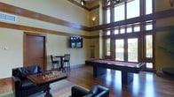This single-story game room is a well-appointed entertainment space defined by its dramatic double-height ceilings and an expansive wall of wood-framed windows and glass doors. The towering windows flood the interior with natural light and offer views of a balcony or terrace with mature trees beyond, creating an open, airy atmosphere despite the richly warm interior palette.

The space features rich hardwood flooring throughout, complemented by warm yellow-toned walls accented with dark wood trim that runs horizontally, adding architectural character and depth. A full-size billiards table with a deep burgundy felt surface serves as the focal centerpiece, accompanied by a mounted cue rack nearby. A wall-mounted flat-screen television provides an additional entertainment option.

A high-top bistro table with bar-height chairs occupies one corner, offering a casual seating vignette, while a comfortable lounge seating area with upholstered chairs anchors another section of the room. A chess set displayed on a low wooden table adds a classic gaming element. Recessed and wall-mounted lighting fixtures complement the abundant natural light, ensuring the space remains well-lit and inviting throughout the day.