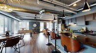 Located at 1430 Whiterock Ave in Waukesha, Wisconsin, this single-floor space showcases a striking industrial-chic aesthetic throughout its open, flowing layout. Exposed concrete ceilings with polished silver ductwork running overhead lend an unmistakable urban character, while rich hardwood-style flooring grounds the space with warmth and contrast.

The kitchen is a standout feature, equipped with dark espresso cabinetry, granite countertops, stainless steel appliances including a full-size refrigerator and range, and a central island with a curved bar overhang fitted with stylish swivel stools. Pendant lighting in bold geometric forms hangs above the island, adding visual drama.

The open-plan layout transitions seamlessly into a comfortable lounge area furnished with a cognac leather sofa, accent chairs, a patterned area rug, and a round wood coffee table. Large windows with horizontal blinds frame views of the surrounding neighborhood and invite generous natural light throughout the day.

A dedicated game area features a foosball table and a rustic high-top table with metal stools positioned in a windowed corner—ideal for casual gatherings. The cohesive slate-blue wall palette ties the entire space together, creating a contemporary, welcoming ambiance suited for both relaxation and social activity.