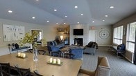 This well-appointed single-floor space offers an open, flowing layout designed for comfort and functionality. The main living area is anchored by a striking floor-to-ceiling stone fireplace, which serves as the room's focal point and is complemented by a wall-mounted flat-screen television above. Recessed lighting is installed throughout the vaulted ceiling, providing consistent, bright illumination across the entire space.

The lounge area features multiple seating arrangements, including plush navy blue sofas and armchairs paired with sleek coffee tables, all set atop a neutral area rug. This creates a warm and inviting atmosphere ideal for gathering and relaxation. Additional seating is tucked into a cozy nook toward the side of the room, offering a more intimate setting.

Adjacent to the lounge, a dining area is furnished with a large rectangular table and upholstered chairs, decorated with elegant candle centerpieces. Decorative greenery and tasteful accents are placed throughout, adding life and warmth to the interior. A large decorative clock adorns one wall, contributing to the space's refined aesthetic. The overall design palette of soft grays, navy blues, and warm neutrals lends the space a cohesive, contemporary character throughout.