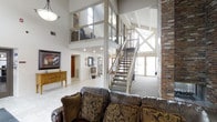 This remarkable space makes an immediate impression with its soaring double-height ceilings and open, airy layout. The main living area is anchored by a rich leather sofa ensemble, complemented by decorative throw pillows that add warmth and texture. A classic wooden sideboard with framed artwork above adds a refined touch to the room.

One of the space's most defining architectural features is the open mezzanine level, accessible via a carpeted staircase with a sturdy metal and wood railing. Large glass-paned panels line the mezzanine's edge, allowing light to flow freely between levels and maintaining a seamless visual connection throughout. Exposed brick columns and walls introduce a rustic, industrial character that contrasts beautifully with the clean white walls and light tile flooring below.

Natural light pours in through expansive glazed entry doors and upper-level windows, creating a bright and welcoming atmosphere throughout. Recessed lighting supplements the natural illumination, ensuring the space is well-lit at all times. Beyond the main living area, additional rooms are visible, suggesting a generous and well-connected floor plan. The overall ambiance is one of open, versatile living, where architectural drama meets everyday comfort.