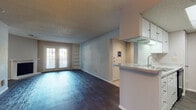 Located at 11710 Parliament St in San Antonio, Texas, this single-story unit offers a well-designed layout with a cohesive blend of comfort and functionality. The open living area is a standout feature, showcasing rich, dark hardwood-style flooring that flows throughout the main spaces. A wood-burning fireplace anchors one wall of the living room, while a set of French doors floods the space with natural light and provides access to an outdoor balcony area. Built-in corner shelving adds both character and practical storage to the room.

The galley-style kitchen is efficiently arranged with white cabinetry, granite-style countertops, a skylight panel for additional illumination, and black appliances including a refrigerator, range, and microwave. A peninsula with a sink opens toward the living area, creating a connected feel between the kitchen and main living space.

The bedrooms offer a quieter retreat, with plush carpeting, generous windows fitted with blinds, and ample natural light. A walk-in closet with built-in shelving and hanging rods provides substantial storage. The textured ceilings are consistent throughout the unit, contributing to its classic residential character. Overall, the space delivers a practical and comfortable living environment in a convenient San Antonio location.