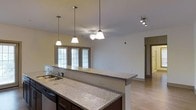 Located at 2945 Midtown Way in Wilmington, North Carolina, this single-story residence offers a thoughtfully designed open-concept layout that seamlessly connects the kitchen and living areas. The kitchen is a standout feature, showcasing expansive granite countertops with speckled detailing, dark-toned cabinetry, and a built-in sink. Elegant pendant lights hang above the kitchen island, providing both task lighting and visual appeal, while a ceiling fan in the adjoining living area ensures year-round comfort. Track lighting adds an additional layer of illumination to the space. Large windows along the exterior wall flood the interior with natural light, creating a bright and welcoming ambiance throughout. Light-toned hardwood-style flooring runs through the open living area, contributing to a clean and contemporary aesthetic. The neutral wall palette enhances the sense of openness, while a hallway visible in the background suggests well-organized private quarters beyond the main living space. Overall, this residence combines practical design with modern finishes to offer a comfortable and functional living environment in a convenient Wilmington location.