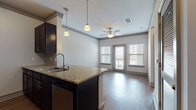 This single-floor residence is designed with an open-concept layout that seamlessly connects the kitchen and living area. The kitchen serves as a central focal point, featuring dark espresso cabinetry, granite countertops, a peninsula with an integrated sink, and stainless steel appliances including a refrigerator, microwave, and range. Pendant lighting above the peninsula adds a refined touch, complementing the tile backsplash and track lighting overhead. The living area extends naturally from the kitchen, showcasing hardwood-style flooring, a ceiling fan, and large windows flanking a glass exterior door that fills the space with natural light and provides outdoor access. A louvered closet door and additional storage are visible near the entryway, adding functional versatility. The bedroom offers plush carpeting, a ceiling fan, crown molding detail, and a generously sized window that brings in ample natural light. The bathroom is equipped with a soaking tub and shower combination featuring tile surround, wood-style flooring, a dark vanity with a granite countertop, and clean, neutral finishes throughout. Overall, the space presents a cohesive, well-appointed aesthetic with consistent finishes across all areas.
