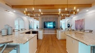 This single-floor community amenity space is designed with a refined coastal-contemporary aesthetic that balances style and functionality throughout. The heart of the space is a striking open-plan kitchen and bar area featuring dual granite-topped islands with white cabinetry, wide-plank hardwood flooring, and dramatic modern chandeliers suspended from exposed wood beam ceilings. A large wall-mounted television anchors a textured feature wall, creating a natural focal point between the islands. Adjacent to the kitchen, a generous lounge area is furnished with plush neutral sofas, layered accent chairs, and gold-toned coffee tables atop woven rugs. Exposed wood beams, recessed lighting, and arched architectural details add warmth and character throughout. A dedicated business center offers multiple workstations along a sleek built-in desk with a textured accent wall and ample natural light. The game room is a bright, airy retreat with large windows, hardwood floors, a full-size pool table, and a shuffleboard table. Intimate lounge nooks are finished with a bold gold geometric accent wall, pendant chandeliers, built-in banquette seating, and cozy lounge chairs — ideal for small gatherings or quiet conversation. Together, these spaces form a cohesive, upscale environment suited for socializing, working, and recreation.