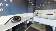 This single-level pet grooming space is thoughtfully designed with both functionality and personality in mind. The room is finished with deep navy blue walls accented by white paw print decals, creating a playful yet purposeful atmosphere. A two-tone design separates the upper wall from a crisp white lower wainscoting, adding a clean, structured aesthetic.

The centerpiece of the space is a raised, white utility wash tub equipped with a wall-mounted sprayer and hose attachment, making pet bathing efficient and accessible. Adjacent to the tub, an open shelving unit provides generous counter space and lower storage, currently stocked with neatly folded towels. A professional-grade pet dryer with a flexible hose rests on the countertop, ready for use.

The flooring features a durable, easy-to-clean surface with yellow safety striping, and a floor drain ensures practical water management. A textured entry mat adds slip resistance near the entrance. Overall, the space is compact yet well-equipped, offering everything needed for a complete and convenient pet grooming experience.