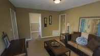 This single-floor residence offers a warm and inviting atmosphere defined by its earth-tone color palette and neutral carpeted flooring throughout. The living area serves as the heart of the home, featuring a comfortable sofa, a dark wood coffee table, and a coordinating side table with a classic lamp. Decorative framed wall art adds a personal touch to the warm golden-tan walls, which are accented by crisp white trim and baseboards. A textured ceiling with flush-mount lighting provides consistent illumination across the space. Doorways from the living area lead to additional rooms, including what appears to be a bedroom and a connected space that may serve as a walk-in closet or secondary room, both maintaining the same cohesive color scheme and carpeted flooring. The layout is thoughtfully designed to maximize flow and connectivity between spaces, creating a seamless and functional living environment. The overall ambiance is cozy and understated, with a neutral foundation that offers a versatile canvas for a variety of interior styles.