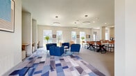The Gibson Clubhouse at 2422 S Carrier Pkwy in Grand Prairie, Texas, is a thoughtfully designed community space that blends modern aesthetics with versatile, social-focused amenities. The single-floor layout offers a generous, open-concept footprint that flows seamlessly between a welcoming lounge area, a dining space, and a dedicated game room. A bold, eye-catching mural branded with "The Gibson" in neon lettering serves as a striking focal point near the entrance, setting the tone for the vibrant atmosphere throughout. The lounge area features plush blue armchairs and neutral sofas arranged around a geometric, multi-toned carpet, all beneath elegant circular pendant lighting that adds a contemporary touch. Natural light pours in through multiple large windows, offering views of the outdoor pool area. The dining and activity zone is anchored by a large ping-pong table surrounded by tan leather and black metal chairs, alongside counter-height bar seating along the walls. A wall-mounted TV and curated photo gallery add personality to the space. Elsewhere, a pool table, potted palms, and a rustic American flag artwork contribute to the clubhouse's eclectic, community-centered character. Recessed lighting and clean white walls throughout create a bright, inviting ambiance ideal for gatherings and recreation.