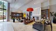This thoughtfully designed single-floor amenity space offers a sophisticated blend of comfort and entertainment across several interconnected zones. The lounge area anchors the space with soaring ceilings, a sleek fireplace framed by built-in shelving, and an eye-catching oversized orange pendant light that adds a bold design statement. Plush velvet sofas, woven accent chairs, and layered area rugs create a warm, inviting atmosphere complemented by curated artwork on the walls.

A bright entrance foyer features floor-to-ceiling glass doors and windows that flood the space with natural light and offer views of a lush outdoor area, likely a pool courtyard lined with palm trees. Sheer white curtains frame the glazing elegantly. Adjacent to the entry, a seating nook with built-in gray banquette booths, wall-mounted televisions, and mustard-yellow accent chairs provides a more intimate setting for smaller gatherings.

A long bar counter with bar stools and a bank of wall-mounted screens serves as a media and social hub, benefiting further from the dramatic double-height glazing nearby. A dedicated game room rounds out the amenities, featuring foosball and shuffleboard tables, colorful pop-art wall pieces, and direct sightlines to the outdoor area, completing this vibrant, multi-functional community space.