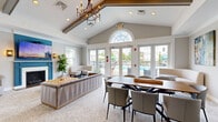 Located at 330 Jacksonville Rd in Warminster, PA, this well-appointed single-floor clubhouse space offers a thoughtfully designed layout comprising an open-plan living and dining room alongside a functional kitchen. The living area is a standout, featuring soaring vaulted ceilings with exposed wood beams and a striking teal-accented fireplace with a wall-mounted television above, creating a warm and inviting focal point. Natural light pours in through multiple windows and a set of glass French doors that open to an outdoor pool area, offering pleasant views and a seamless indoor-outdoor connection. A large arched transom window further enhances the airy atmosphere. The space is finished with neutral-toned patterned carpeting throughout the living and dining areas, complementing the light gray walls and white trim. The dining area accommodates a generous wooden table with upholstered chairs and features colorful abstract wall art, adding a creative touch. An interior window connects the dining area to adjacent spaces, providing visual openness. The kitchen is equipped with rich warm-toned wood cabinetry, light countertops, stainless steel appliances including a full-size refrigerator and built-in microwave, and tile flooring with crown molding detail. Together, these spaces create a versatile, comfortable, and well-finished communal environment.