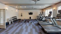Located at 350 Bradley Park Lane in Cumming, Georgia, this single-floor fitness center offers a comprehensive and thoughtfully arranged workout facility. The space is designed with a clean, modern aesthetic, featuring neutral-toned walls, recessed lighting, wall-mounted sconces, and multi-toned striped carpet tiles that define distinct workout zones throughout the room.

The cardio area is well-stocked with Matrix-brand treadmills, ellipticals, and upright bikes, positioned along the perimeter to take advantage of large windows fitted with horizontal blinds. These windows allow ample natural light to filter into the space and provide views of the surrounding outdoor area, including a glimpse of a pool. Ceiling fans overhead contribute to airflow and comfort during workouts.

The strength training section includes a multi-station cable and weight machine, along with a dedicated free weights area featuring a tiered dumbbell rack, medicine balls, stability balls, and a flat workout bench. A wall-mounted television is positioned for easy viewing from the cardio equipment zone.

The open floor plan allows for flexible use of the central space, while a glass-paneled door provides access to an adjacent area. Informational signage is prominently displayed near the entrance, contributing to an organized and user-friendly environment.