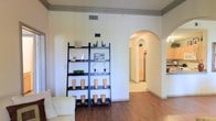 This single-floor residence presents a clean, well-organized layout with a warm and inviting ambiance. The living area showcases rich hardwood flooring that flows seamlessly throughout the space, complemented by crisp white walls and crown molding that lend a classic, polished character to the interior. A standout architectural feature is the graceful arched doorway that connects the main living area to the adjacent spaces, adding an elegant and timeless design element. The living room is furnished with a neutral-toned sofa and accented by a sleek, open metal shelving unit displaying a curated arrangement of decorative objects, adding both function and visual interest to the room. The high ceilings enhance the sense of openness and allow light to circulate freely, creating a bright and pleasant atmosphere. Through the arched passage, the space opens to a hallway leading toward what appears to be a well-lit kitchen, featuring light-toned cabinetry that reinforces the home's consistently bright and airy aesthetic. Ventilation is thoughtfully integrated with ceiling vents maintaining comfort throughout. Overall, the space balances architectural character with practical, livable design in a cohesive and well-maintained interior.