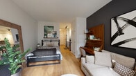 This single-floor studio is designed to maximize every inch of its open layout, seamlessly blending the sleeping, living, and working areas into one cohesive space. Warm hardwood-style flooring runs throughout, lending continuity and a welcoming feel. The sleeping area is anchored by a platform bed, accompanied by a wall-mounted shelf for display, while a bold dark accent wall adds depth and visual contrast to the room.

A dedicated workspace features a warm-toned slatted wood panel backdrop with floating shelves, paired with a mid-century style desk — a distinctive design element that elevates the studio's character. The living area benefits from large windows that invite generous natural light and urban views, creating an airy atmosphere.

The kitchen is efficiently outfitted with white shaker cabinetry, quartz countertops, a white subway tile backsplash, stainless steel appliances including an electric range and over-the-range microwave, and a matte black faucet. The bathroom continues the modern aesthetic with a white vanity featuring black hardware, a framed mirror, quartz countertop, and classic dark hexagonal penny tile flooring. A stacked in-unit washer and dryer adds everyday convenience.