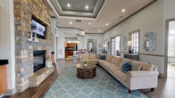 Located at 1475 Benton Blvd in Pooler, Georgia, this single-floor community clubhouse offers a thoughtfully designed gathering space with upscale finishes throughout. The expansive main living area is anchored by a dramatic floor-to-ceiling stacked stone fireplace with a wall-mounted television, complemented by a coffered tray ceiling with recessed lighting and rich hardwood-style flooring. A generously sized sectional sofa and patterned area rug define the seating zone, creating a welcoming atmosphere.

Adjacent to the main lounge, a cozy dining nook bathes in natural light from large multi-pane windows and features an elegant pendant light fixture overhead. The kitchen area includes warm wood cabinetry, dark countertops, a central island with bar seating, and a crystal chandelier, offering both functionality and style.

A secondary sitting room provides a quieter retreat with tile flooring, abundant natural light, and comfortable seating. Built-in display cabinetry flanks the fireplace wall, adding architectural character throughout.

The screened outdoor lounge is a standout feature, wrapped in floor-to-ceiling windows that frame views of the surrounding community and pool area. Outfitted with vibrant teal sectional seating, ceiling fans, and tile flooring, this space is ideal for relaxed social gatherings year-round.