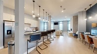 The Walden Lakes clubhouse at 3820 Codet Rd in Columbus, Ohio, is a thoughtfully designed single-floor community space that combines functionality with contemporary style. The layout opens from a spacious entry corridor finished with light hardwood-style flooring and soft gray walls, guiding visitors toward distinct functional zones throughout the facility.

At the heart of the space is a prominent reception desk bearing the Walden Lakes name, set beneath gleaming pendant lights and accented by a textured quilted feature wall and mounted display screen. Adjacent to reception, a well-equipped kitchenette offers white shaker cabinetry, stainless steel appliances, dark countertops, and a long breakfast bar lined with industrial-style stools beneath a row of globe pendant lights—ideal for casual gatherings.

The lounge area features bold navy-blue walls, a wall-mounted television, and generously sized seating arranged around a wood coffee table, creating an inviting atmosphere for socializing. Natural light filters through large, curtain-framed windows throughout. A dedicated business center with computer workstations provides a productive workspace. Additional flex rooms with neutral tones and natural light offer versatile utility. Together, these elements create a polished, welcoming communal environment.
