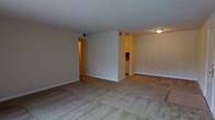 This single-floor residential space offers a straightforward and functional layout designed for comfortable everyday living. The main living area is spacious and open, featuring wall-to-wall carpet flooring in a neutral tone that adds warmth underfoot. The walls are painted in a clean, light beige palette, creating a bright and welcoming atmosphere throughout.

A central partial wall divides the living space, lending subtle definition to the layout while maintaining an open feel. Two pass-through openings on either side of this dividing wall provide seamless access to adjoining areas. To one side, a glimpse into the kitchen reveals cabinetry with a warm wood finish and white lower cabinets, hinting at a practical and organized cooking space. The opposite opening leads into a hallway corridor, suggesting access to additional rooms deeper within the unit.

Overhead lighting is provided by a flush-mount ceiling fixture, complementing the recessed ceiling vents that indicate a central HVAC system for year-round climate control. Crisp white baseboards run along the perimeter of the rooms, adding a clean finishing touch to the overall interior. The single-floor layout ensures easy navigation throughout the space, making it an accessible and efficient home.