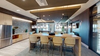 This thoughtfully designed single-floor amenity space offers a sophisticated blend of entertainment, relaxation, and social gathering areas, all unified by a cohesive contemporary aesthetic. The open lounge area anchors the space with a curved sofa, a wall-mounted TV set into rich wood paneling, and plush carpeting, bathed in natural light streaming through generously sized windows. Adjacent to the lounge, a spacious communal dining area features a large wooden table with upholstered bar-height chairs beneath a striking row of industrial-style chain-hung pendant lights, complemented by a wet bar with light wood cabinetry, marble-look countertops, and a herringbone tile backsplash. Two billiards rooms provide dedicated entertainment areas, each featuring a full-size pool table, carpeted flooring, cue racks, and ample natural light from large windows. A private screening room offers plush lounge chairs, teal accent ceilings with dark wood paneling, a wall-mounted display screen, and dramatic drape detailing for an intimate theater experience. A marble-topped nook with a textured slate tile accent wall and an outdoor view adds a quiet retreat. Wide-plank wood-look flooring runs through the corridors, connecting all areas with a clean, modern flow throughout.