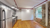 Located at 111 N Marietta Pkwy NE in Marietta, GA, this one-bedroom, one-bathroom unit offers a compelling blend of historic character and modern updates within a thoughtfully converted building. The open-concept living, dining, and kitchen area serves as the heart of the home, showcasing wide-plank hardwood floors, exposed red overhead pipework, and original stone masonry walls flanking large windows that flood the space with natural light and views of surrounding greenery. The kitchen is outfitted with stainless steel appliances — including a gas range, microwave, and refrigerator — gray cabinetry, and pendant industrial lighting. The bedroom features a distinctive black cast-iron column, dark flooring, exposed red pipes overhead, and a ceiling fan, reinforcing the unit's loft-like industrial aesthetic. A long corridor with dark flooring and exposed ceiling elements connects the spaces efficiently. The bathroom includes a full soaking tub and shower combo with warm earth-toned tile surround and dark oil-rubbed bronze fixtures. Additional conveniences include a dedicated laundry area and a closet for storage. The unit's raw architectural details, including original stonework and structural columns, lend it a one-of-a-kind character rooted in the building's history.