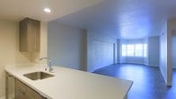 This single-floor space showcases an open-concept design that seamlessly connects the kitchen and living area, creating a cohesive and functional layout. The kitchen is equipped with a sleek, light-colored countertop that extends generously, providing ample workspace and a casual bar-style overhang. A stainless steel sink with a modern arc-neck faucet is integrated into the countertop, adding a contemporary touch. Recessed lighting illuminates the kitchen area from above.

The adjoining living space is defined by neutral-toned walls and features a notable decorative accent wall clad in small mosaic-style tiles, serving as a distinctive focal point. Large windows along the far wall allow natural light to filter into the living area, brightening the space and contributing to an airy atmosphere. The flooring transitions between the two areas and appears to be a smooth, dark material in the living space. The textured ceiling throughout adds subtle visual interest. Overall, the space presents a clean, modern aesthetic well-suited for everyday living.