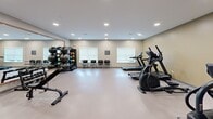 Located at 6822 Shannon Parkway in Union City, Georgia, this fitness center offers a well-appointed exercise space designed to accommodate a range of workout needs. The single-room layout provides a generous open floor plan with durable, light-toned flooring that allows for easy movement between equipment stations.

The cardio section is equipped with multiple treadmills and elliptical machines, positioned to take advantage of natural light from the roller-shaded windows along one wall. A multi-functional cable machine and weight bench serve the strength training area, while a dedicated free weights station features an organized rack of medicine balls, exercise balls, and dumbbells for varied training options.

Large wall-mounted mirrors along one side of the room enhance the sense of space and allow users to monitor their form during workouts. Recessed ceiling lighting provides consistent, even illumination throughout. The neutral color palette — combining warm gold and soft gray tones on the walls — creates a clean, motivating atmosphere.

Seating is available near the entrance, which features a wood-framed glass door and an emergency exit sign overhead. A stainless steel waste receptacle and decorative wall art add functional and aesthetic finishing touches to the space. The overall design is practical, organized, and well-suited for a focused fitness experience.