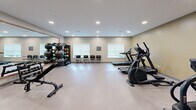 Located at 6822 Shannon Parkway in Union City, Georgia, this fitness center offers a well-appointed exercise space designed to accommodate a range of workout needs. The single-room layout provides a generous open floor plan with durable, light-toned flooring that allows for easy movement between equipment stations.

The cardio section is equipped with multiple treadmills and elliptical machines, positioned to take advantage of natural light from the roller-shaded windows along one wall. A multi-functional cable machine and weight bench serve the strength training area, while a dedicated free weights station features an organized rack of medicine balls, exercise balls, and dumbbells for varied training options.

Large wall-mounted mirrors along one side of the room enhance the sense of space and allow users to monitor their form during workouts. Recessed ceiling lighting provides consistent, even illumination throughout. The neutral color palette — combining warm gold and soft gray tones on the walls — creates a clean, motivating atmosphere.

Seating is available near the entrance, which features a wood-framed glass door and an emergency exit sign overhead. A stainless steel waste receptacle and decorative wall art add functional and aesthetic finishing touches to the space. The overall design is practical, organized, and well-suited for a focused fitness experience.