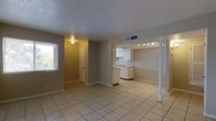 This single-story space features an open, flowing layout that connects the main living area to the kitchen, creating a functional and cohesive floor plan. Light beige ceramic tile flooring extends throughout, providing a clean, low-maintenance surface that unifies the space visually.

The living area is finished with neutral gray-toned walls and receives natural light from a window on the side wall, fitted with horizontal blinds. The textured ceiling adds subtle dimension, while ceiling-mounted HVAC vents ensure efficient climate control throughout.

The kitchen is positioned toward the rear and is defined by white cabinetry, an under-cabinet range hood, and a sink along the counter. Track lighting above the kitchen provides focused illumination. A decorative twisted column and a partial half-wall visually separate an adjacent area, adding an architectural detail that breaks up the open floor plan without fully enclosing the space.

A hallway near the kitchen leads to additional rooms, with a door visible at the end suggesting access to private spaces. The overall ambiance is clean, neutral, and practical, offering a straightforward layout suited for everyday living.