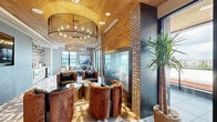 Located along Waterside Drive in Nashville, Tennessee, this thoughtfully designed lounge space blends industrial character with upscale finishes across a single open floor. The living area makes a strong architectural impression with its warm wood-paneled ceiling, bold geometric-patterned tile flooring, and a striking exposed brick accent wall that anchors a glass-enclosed fireplace. Two dramatic drum-style chandeliers provide a focal point overhead, casting warm ambient light throughout the room. Plush barrel chairs in rich cognac leather invite comfortable seating, complemented by sleek metallic accent tables that add a contemporary touch. A built-in kitchenette along one wall features natural wood cabinetry, open industrial shelving against a brick backdrop, a matte black faucet, and integrated appliances, offering functional convenience within the lounge setting. Floor-to-ceiling sliding glass doors flood the interior with natural light and open directly onto a generous wraparound balcony. The outdoor terrace, finished with large-format tile and a wood-clad ceiling with ceiling fan, offers breathtaking elevated views of the Nashville skyline, the Cumberland River, and the surrounding cityscape — making it a standout feature of this refined urban space.