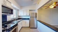 This single-floor residence offers a well-organized layout with distinct living, sleeping, and kitchen areas. The kitchen is a standout feature, designed with white shaker-style cabinetry, dark countertops, and light wood-look flooring. Stainless steel appliances, including a refrigerator, electric range with oven, and over-the-range microwave, provide a modern, functional workspace. A window above the sink allows natural light to brighten the area, while a pass-through counter connects the kitchen to the living space. The living area is carpeted and generously proportioned, with a ceiling fan overhead and direct sightlines into the kitchen. A sliding glass door opens to an outdoor area, offering views of surrounding greenery. The bedrooms are carpeted and naturally lit through windows fitted with vertical blinds, providing a quiet, comfortable atmosphere. The bathroom is clean and well-appointed, featuring a tiled tub-shower combination, a white vanity with dark countertop, dual medicine cabinet mirrors, and light wood-look flooring. Throughout the space, neutral wall tones and consistent finishes create a cohesive, move-in-ready environment.