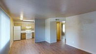 This single-floor residence offers a straightforward, functional layout designed for comfortable everyday living. The open-concept design seamlessly connects the kitchen and living area, creating a cohesive and practical flow throughout the main space. The kitchen is equipped with light wood cabinetry, a white electric range, and an under-cabinet range hood, providing ample storage and workspace. A built-in overhead fluorescent light fixture ensures the kitchen is well-lit for meal preparation. Warm hardwood-style flooring runs continuously throughout the space, adding a sense of warmth and visual consistency. The neutral light gray walls create a clean, versatile backdrop that complements the natural tones of the flooring and cabinetry. A hallway visible from the main living area leads to additional rooms, with a window at the far end drawing in natural light and brightening the interior corridor. Ceiling-mounted HVAC vents are present throughout, ensuring consistent climate control. The overall ambiance is clean, bright, and move-in ready, offering a practical living environment with a simple, uncluttered aesthetic well-suited for a variety of living arrangements.