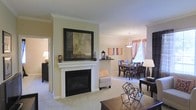 This single-floor residence offers a well-appointed open-concept layout that seamlessly connects the living and dining areas, creating a sense of spaciousness and flow. The living room serves as a central focal point, anchored by a classic white-mantled fireplace with a sleek black surround, above which hangs a framed piece of contemporary artwork. Soft, neutral tones on the walls are complemented by plush carpeted flooring, lending the space a warm and inviting ambiance. Elegant crown molding runs throughout, adding a refined architectural detail that elevates the overall aesthetic. The dining area is defined by a pendant light fixture that casts a warm glow over a dark wood dining table and matching chairs, set against a backdrop of draped windows that allow natural light to filter into the room. Warm-toned curtains frame the windows, softening the space and adding a touch of color. A partial dividing wall subtly separates the two areas while maintaining an open and airy feel. The cohesive design, thoughtful lighting, and classic architectural details combine to create a comfortable and elegantly understated living environment.