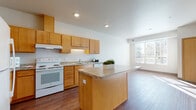 This single-floor unit offers a well-organized, open layout that seamlessly connects the kitchen and living areas. The kitchen is a standout feature, equipped with warm honey-toned wood cabinetry providing ample upper and lower storage, light granite-style countertops, and white appliances including a range, dishwasher, refrigerator, and microwave. A central island with matching countertop serves as both a prep area and a casual gathering space. Wood-look hard flooring extends throughout the kitchen and living area, adding warmth and continuity to the open floor plan. A generously sized window in the living area allows abundant natural light to pour in, offering views of the surrounding outdoor greenery.

The bedroom is carpeted for comfort and bathed in natural light from a large window. It includes access to a walk-in closet fitted with wire shelving, providing practical storage solutions. The bathroom features a single-sink vanity with wood cabinetry and a large mirror, complemented by slate-style tile flooring and accessibility grab bars. Neutral wall tones and clean finishes throughout create a cohesive, calm ambiance across the entire unit.